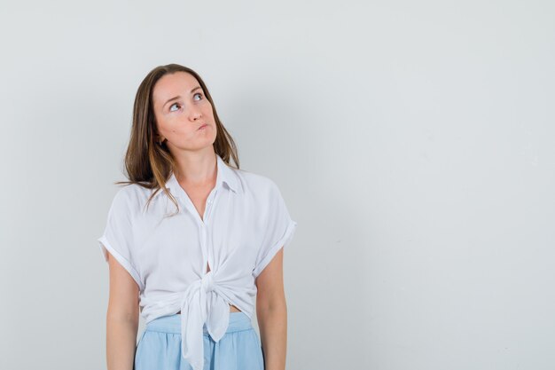 Foto gratuita giovane donna in camicetta bianca e gonna azzurra in piedi dritta e pensando a qualcosa e guardando pensierosa