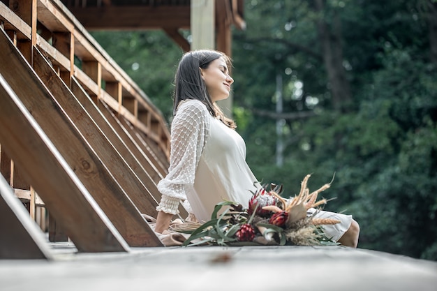 하얀 드레스를 입은 젊은 여성이 이국적인 꽃 꽃다발을 들고 나무 다리에 앉아 있습니다.