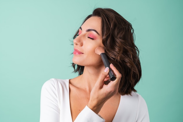 Young woman in a white evening dress on light green with bright pink makeup holding a blush brush