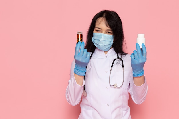 young woman in white medical suit blue gloves blue protective mask holding cans with pills on pink