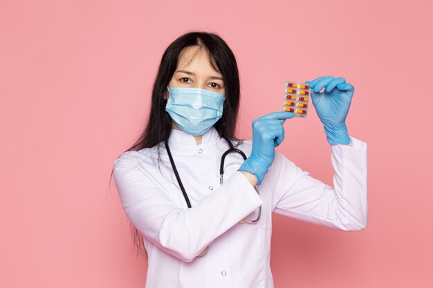 ピンクの多色の錠剤を保持している白い医療スーツ青い手袋青い防護マスクの若い女性