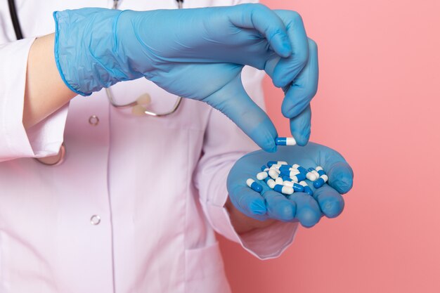 young woman in white medical suit blue gloves blue protective mask holding pills on pink