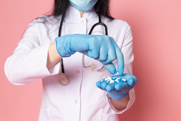 young woman in white medical suit blue gloves blue protective mask holding pills on pink