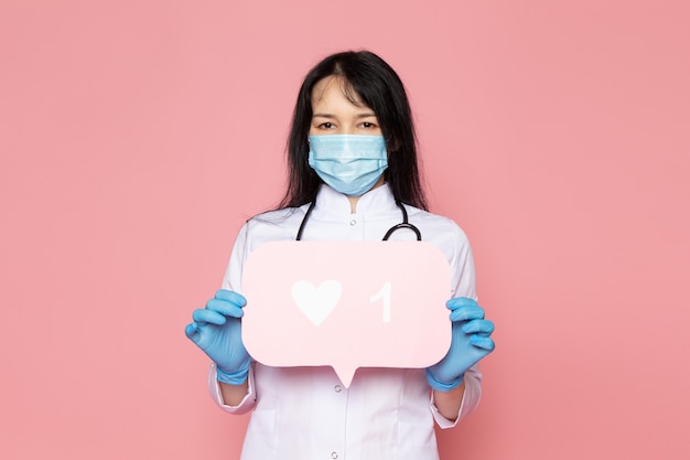 young woman in white medical suit blue gloves blue protective mask with stethoscope holding blank sign on pink