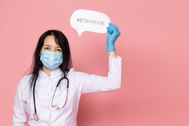 young woman in white medical suit blue gloves blue protective mask with stethoscope holding stayhome hashtag on pink