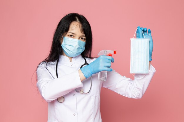 Free photo young woman in white medical suit blue gloves blue protective mask with stethoscope on pink