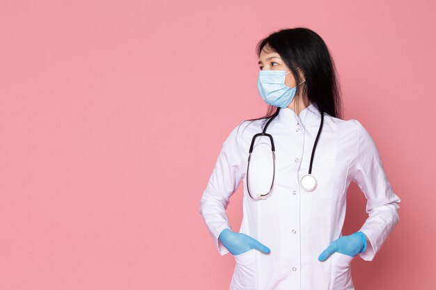 young woman in white medical suit blue gloves blue protective mask with stethoscope on pink