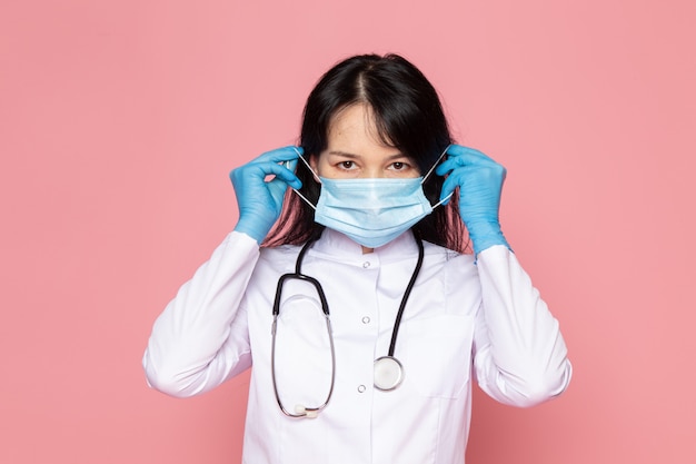 young woman in white medical suit blue gloves blue protective mask with stethoscope on pink