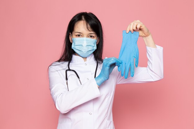 молодая женщина в белом медицинском костюме синие перчатки голубая защитная маска со стетоскопом на розовом