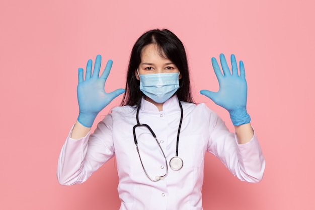 young woman in white medical suit blue gloves blue protective mask with stethoscope on pink