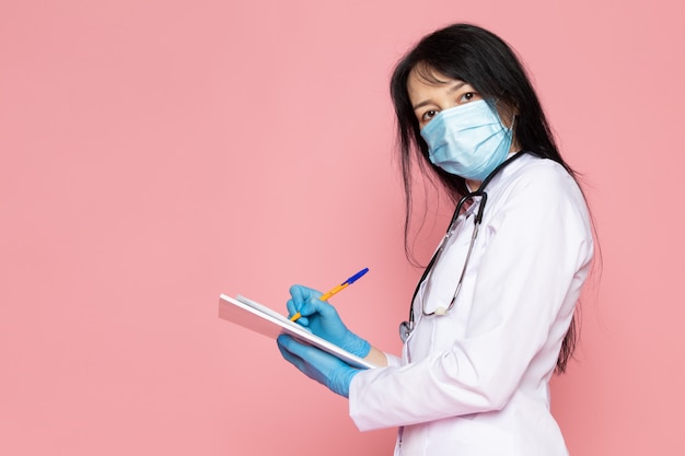 Молодая женщина в белом медицинском костюме синие перчатки синие защитные маски с стетоскопом заметок на розовом