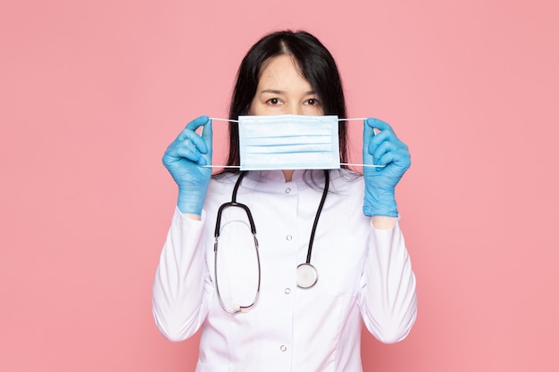 young woman in white medical suit blue gloves white protective mask with stethoscope on pink