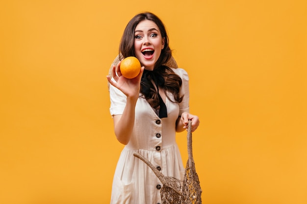 Young woman in white outfit with black bow around her neck gets orange from string bag.