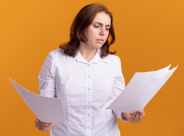 Young woman in white shirt holding blank pages looking at them confused standing over orange wall