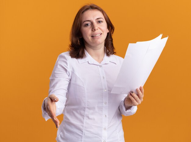 Young woman in white shirt holding blank pages offering hand looking at front smiling greeting standing over orange wall