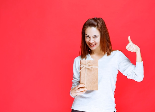 Young woman in white shirt holding a cardboard gift box and showing positive hand sign