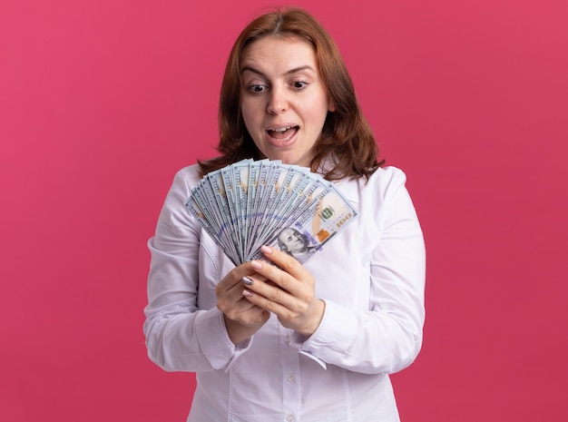 Young woman in white shirt holding cash looking at money happy and excited standing over pink wall