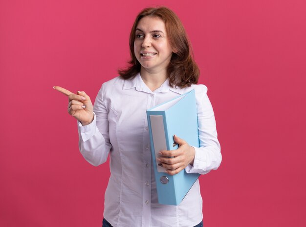Young woman in white shirt holding folder looking aside pointing with index finger to the side smiling cheerfully standing over pink wall