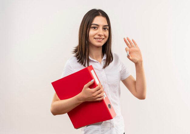 Young woman in white shirt holding folder looking to the front showing ok sign smiling standing over white wall