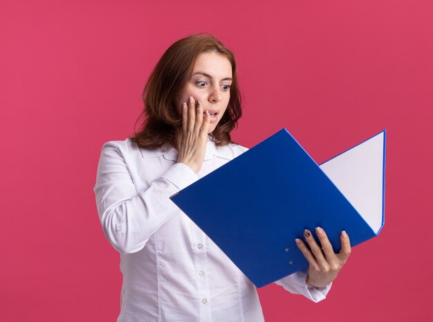 Young woman in white shirt holding folder looking at it amazed and surprised standing over pink wall