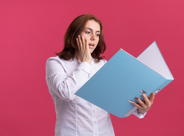 Young woman in white shirt holding folder looking at it amazed and surprised standing over pink wall