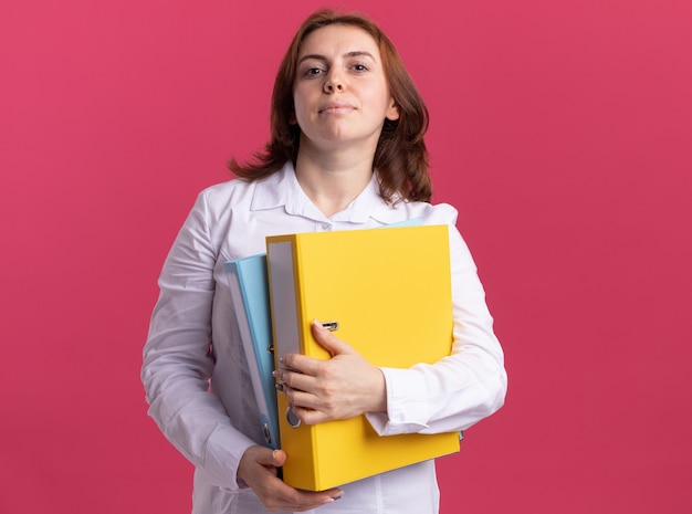 Young woman in white shirt holding folders looking at front with serious confident expression standing over pink wall