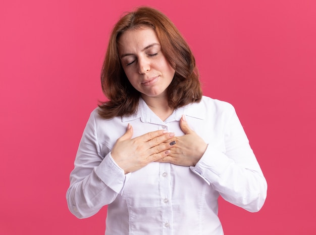 Free photo young woman in white shirt holding hands on her chest feeling thankful standing over pink wall