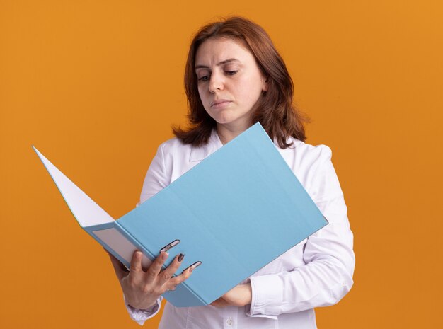 Young woman in white shirt holding open folder looking at it with serious face standing over orange wall