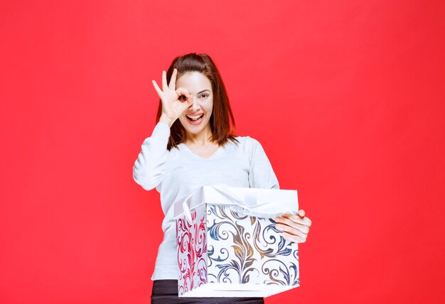 Молодая женщина в белой рубашке держит подарочную коробку с принтом и показывает знак рукой