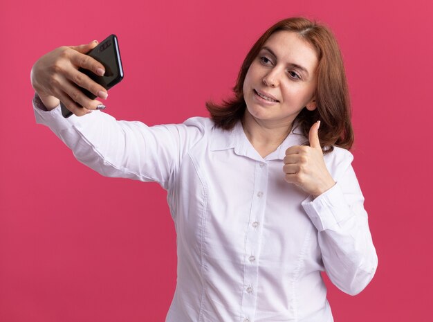 ピンクの壁の上に立って親指を見せて自分撮り笑顔をしているスマートフォンを保持している白いシャツの若い女性