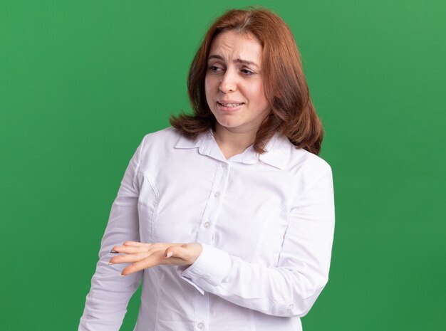 Young woman in white shirt looking aside raising hand in displeasure and indignation standing over green wall