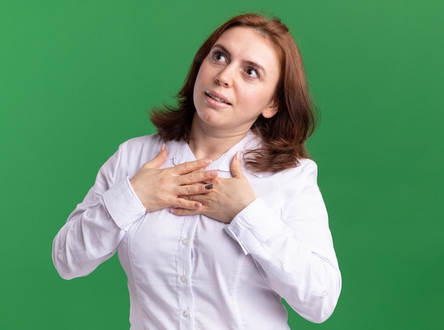 Foto gratuita giovane donna in camicia bianca che osserva da parte con hnads sul petto che si sente grata in piedi oltre il muro verde