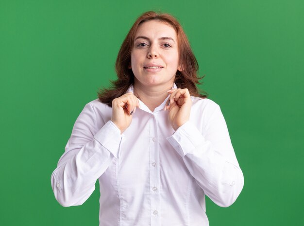 녹색 벽 위에 서있는 그녀의 칼라를 고정하는 자신감을 찾고 흰 셔츠에 젊은 여자
