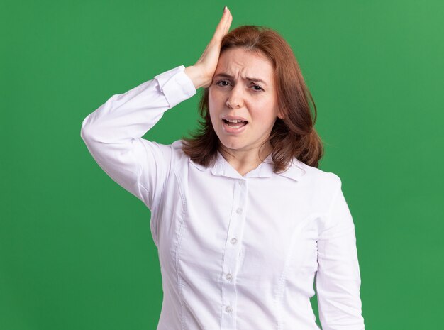 Young woman in white shirt looking confused at front with hand on her head for mistake standing over green wall