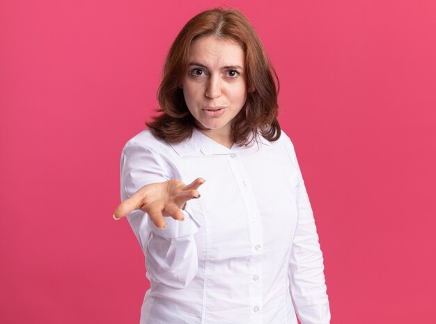 Young woman in white shirt looking at front being displeased with arm out standing over pink wall