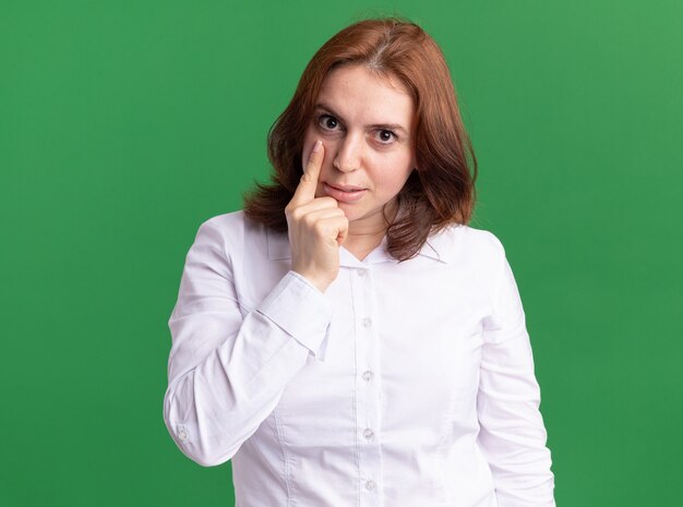 Young woman in white shirt looking at front pointing with index finger at her eye standing over green wall