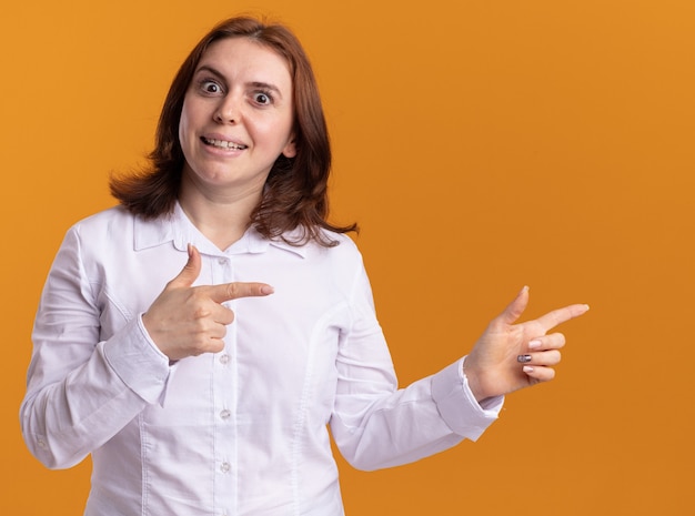 Young woman in white shirt looking at front smiling cheerfully pointing with index fingers to the side standing over orange wall