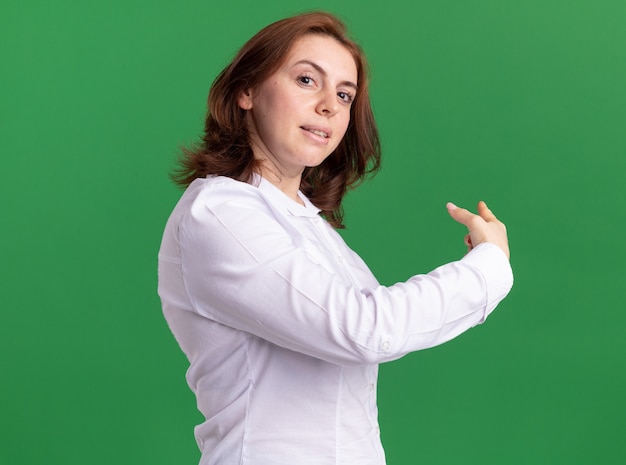 Young woman in white shirt looking at front smiling confident pointing back standing over green wall