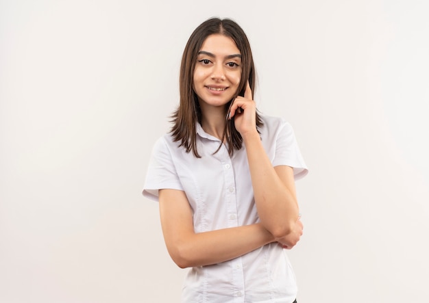 Young woman in white shirt looking to the front smiling confident standing over white wall