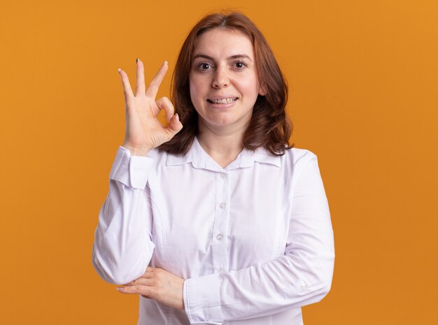 Молодая женщина в белой рубашке, глядя на фронт, смущенно улыбаясь, показывает знак ОК, стоящий над оранжевой стеной