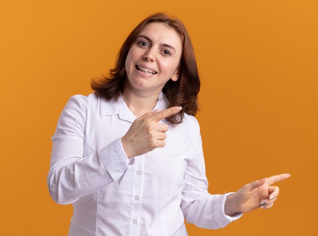 Young woman in white shirt looking at front smiling happy and positive pointing with index fingers to the side standing over orange wall