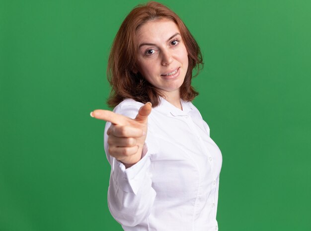 Young woman in white shirt looking at front smiling pointing with index finger at front standing over green wall