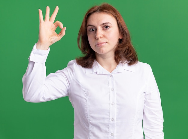 Young woman in white shirt looking at front smiling showing ok sign standing over green wall