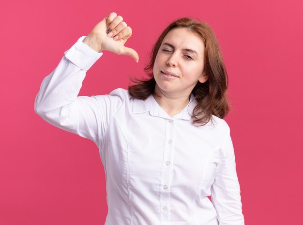 흰 셔츠에 젊은 여자가 웃고 분홍색 벽 위에 엄지 손가락으로 자신을 가리키는 윙크를보고