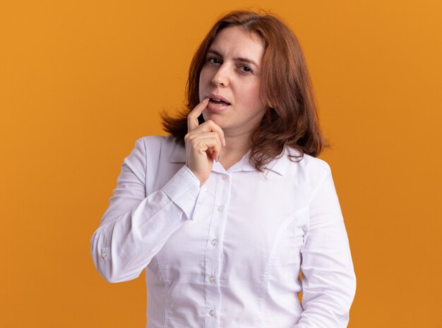 Young woman in white shirt looking at front with pensive expression thinking standing over orange wall