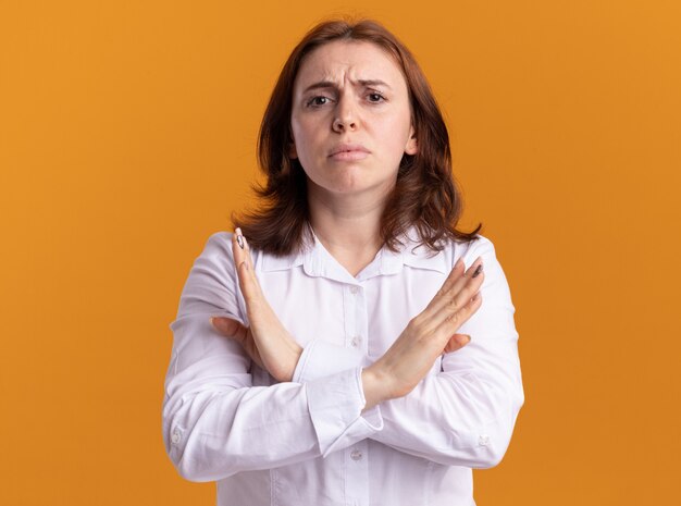 Young woman in white shirt looking at front with serious face making stop gesture crossing hands standing over orange wall