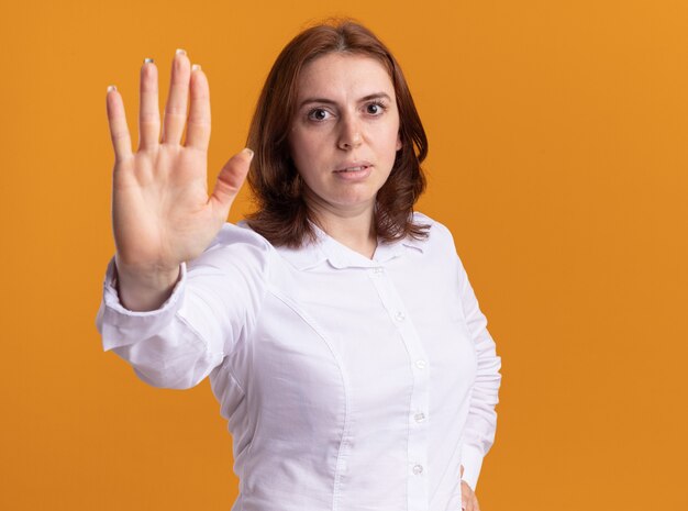 Young woman in white shirt looking at front with serious face making stop gesture with hand standing over orange wall