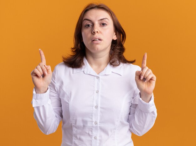 Young woman in white shirt looking at front with serious face pointing with index fingers up standing over orange wall