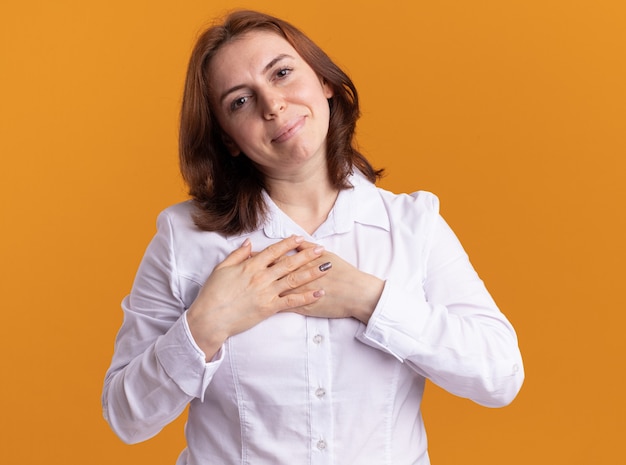 Foto gratuita giovane donna in camicia bianca guardando la parte anteriore con il sorriso sul viso tenendosi per mano sul petto sensazione grata in piedi oltre il muro arancione