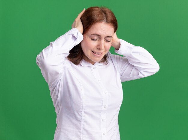 Young woman in white shirt looking unwell touching her head suffering from headache standing over green wall